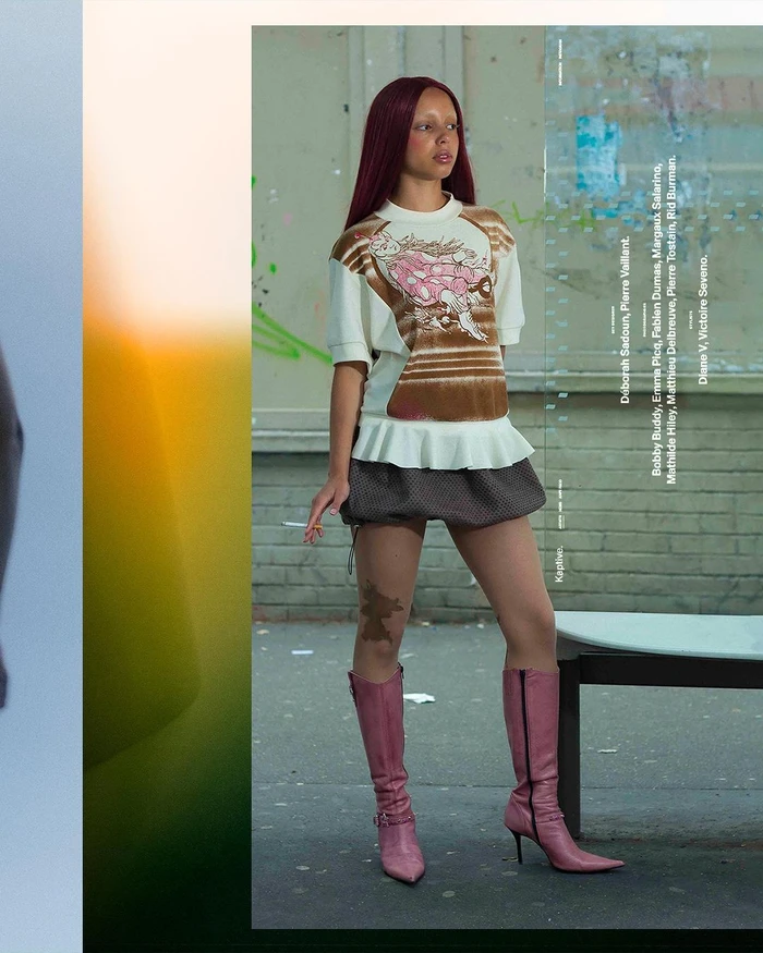 Model with long dark red hair wearing a graphic sweater, skirt, and pink boots standing outdoors.