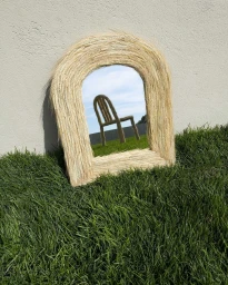 An arched mirror with a woven straw frame rests on vibrant green grass against a textured concrete wall, reflecting a wooden chair outdoors.