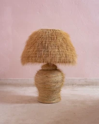 Textured woven straw table lamp with a fringed shade centered against a pale pink wall and floor in Paris