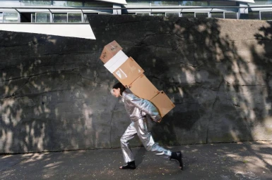 Person in silver metallic jacket and trousers carrying a tall stack of cardboard boxes outdoors