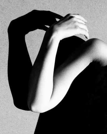 Black and white photograph showcases a woman's arms and shoulder, bathed in shadow and light, creating a dramatic, minimalist composition.