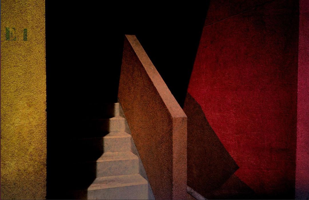 Abstract architectural image with concrete stairs, vivid red wall, textural yellow wall, and strong light and shadow patterns.