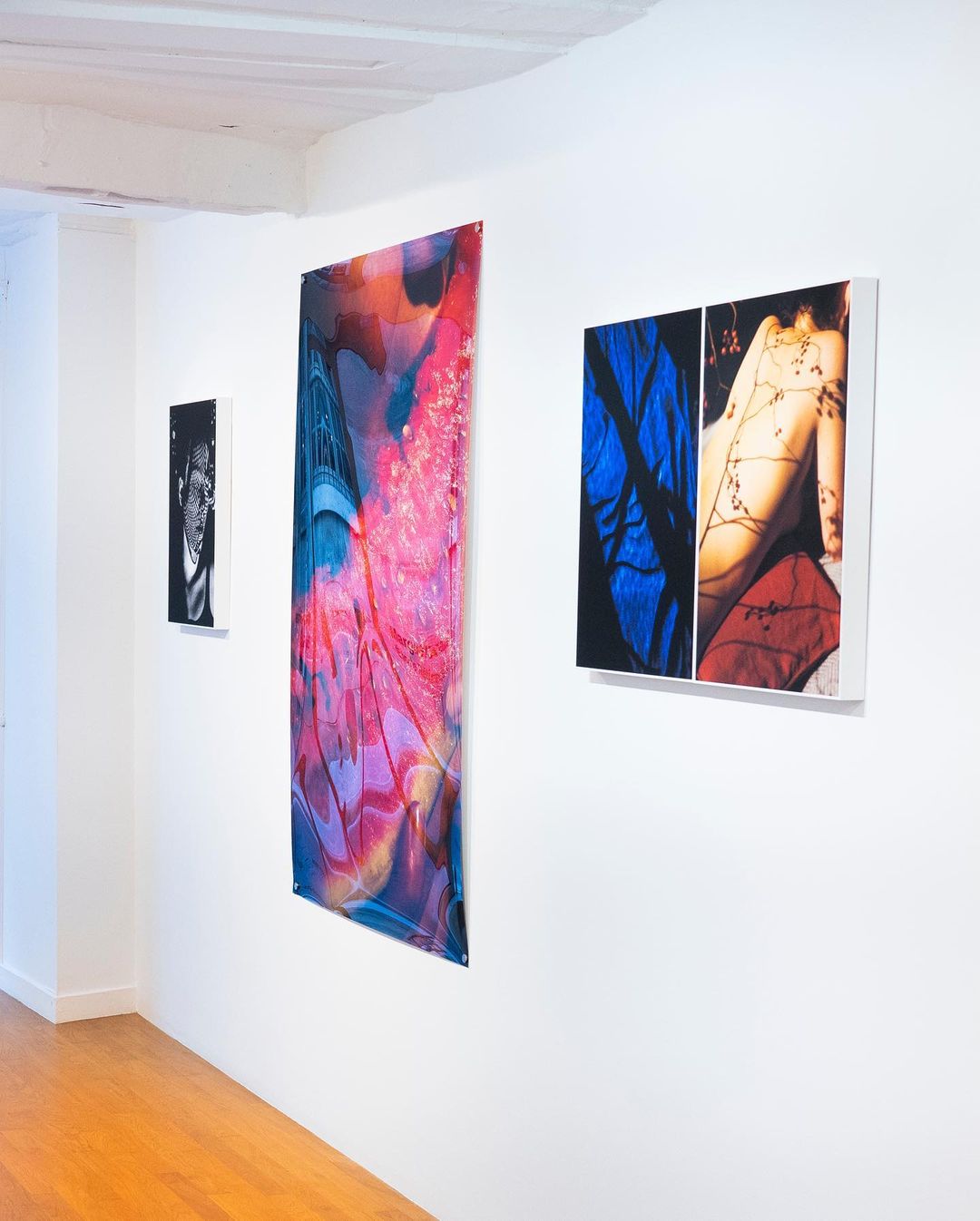 A photo of three artworks hanging on a white wall inside an art gallery. A polished wood floor is also visible.