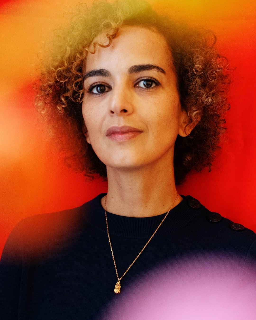 Portrait of an adult female with curly hair in a black top against a blurred red, orange, and pink background. She wears a gold necklace.