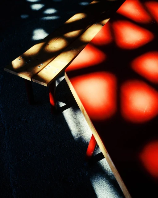 Sunlight creates geometric shadows on a wooden and red bench, highlighting textures and patterns on the ground in an indoor setting.