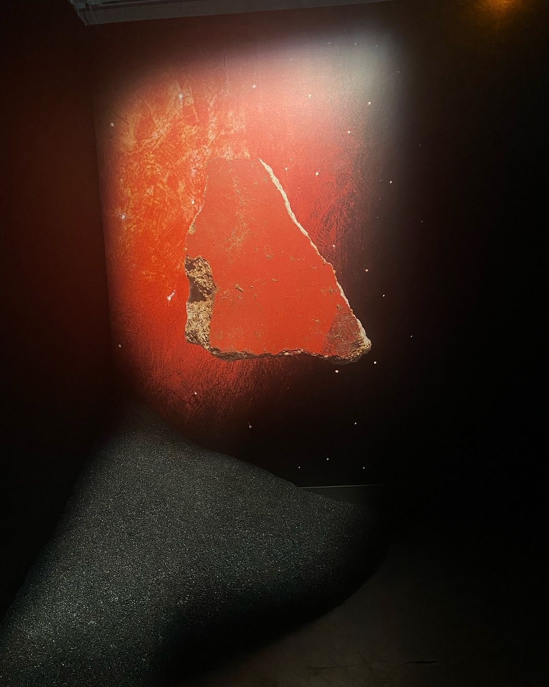 Abstract image of a damaged, red wall with a large missing section revealing rough brown material underneath, against a dark background.