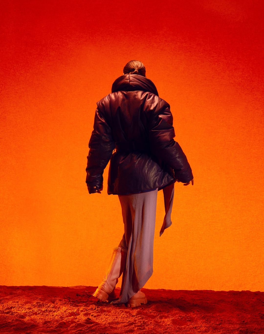 Woman in a black puffer jacket and flowing dress stands in a vivid orange studio, creating a striking fashion and lifestyle image.
