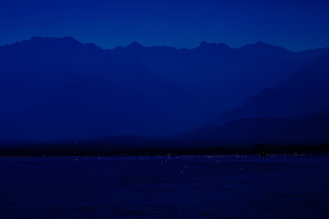Blue monochromatic landscape featuring a tranquil body of water and a majestic mountain range against a deep blue sky at night.