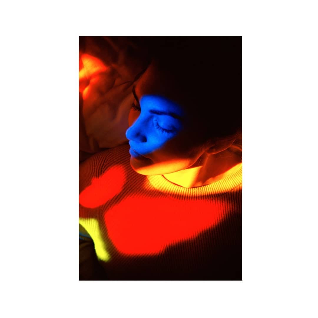 Close-up of a person's face with eyes closed, illuminated by red, yellow, and blue light patterns in a dark indoor setting.