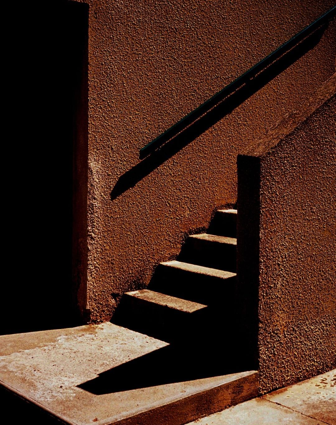 Outdoor staircase made of concrete and stucco with a dark metal handrail, photographed in strong sunlight creating bold shadows.