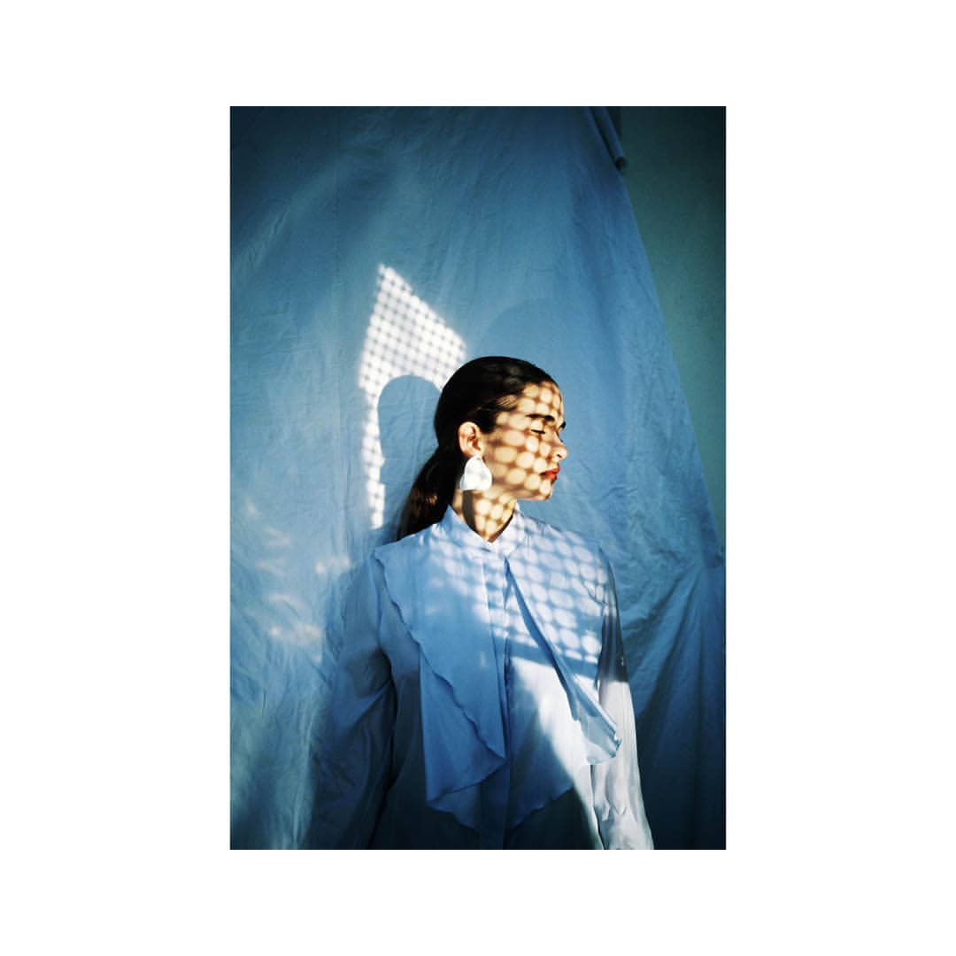 A young woman in a light blue shirt, facing right, with sunlight casting a patterned shadow on her face, against a draped fabric backdrop.