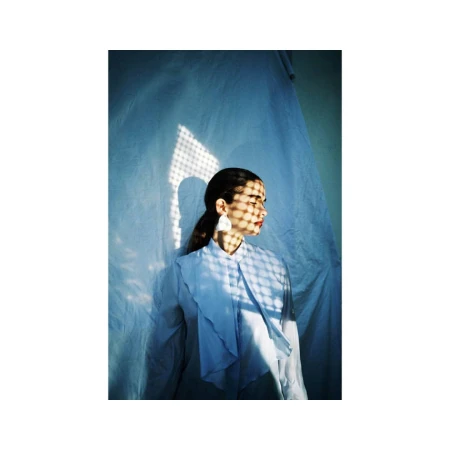 A young woman in a light blue shirt, facing right, with sunlight casting a patterned shadow on her face, against a draped fabric backdrop.