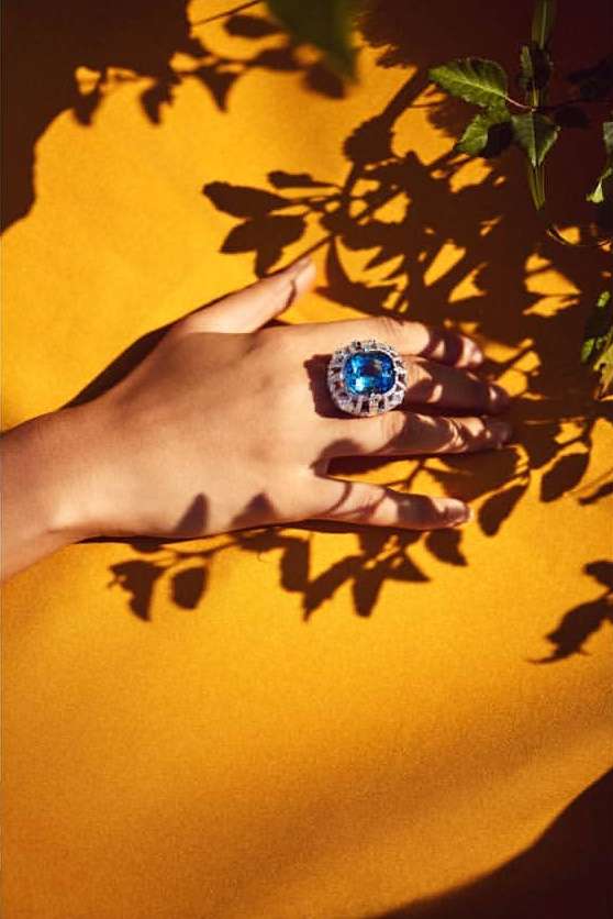 Close-up of a person's hand wearing a blue gemstone ring on a yellow background with leaf shadows adding depth and texture.