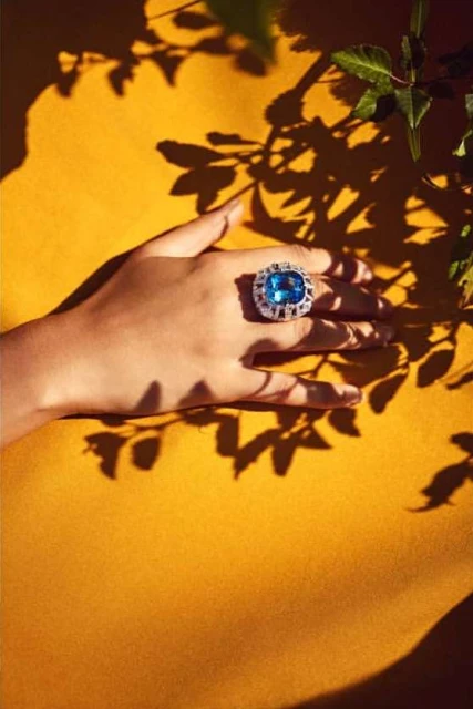 Close-up of a person's hand wearing a blue gemstone ring on a yellow background with leaf shadows adding depth and texture.