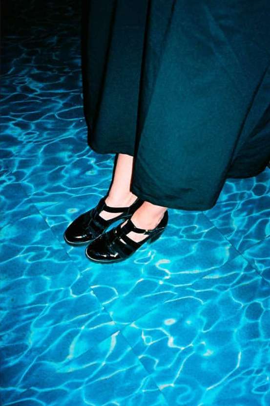 Close-up of feet in black Mary Jane shoes wearing dark trousers standing by blue water caustics.