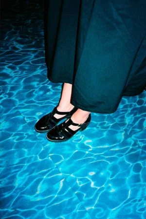 Close-up of feet in black Mary Jane shoes wearing dark trousers standing by blue water caustics.
