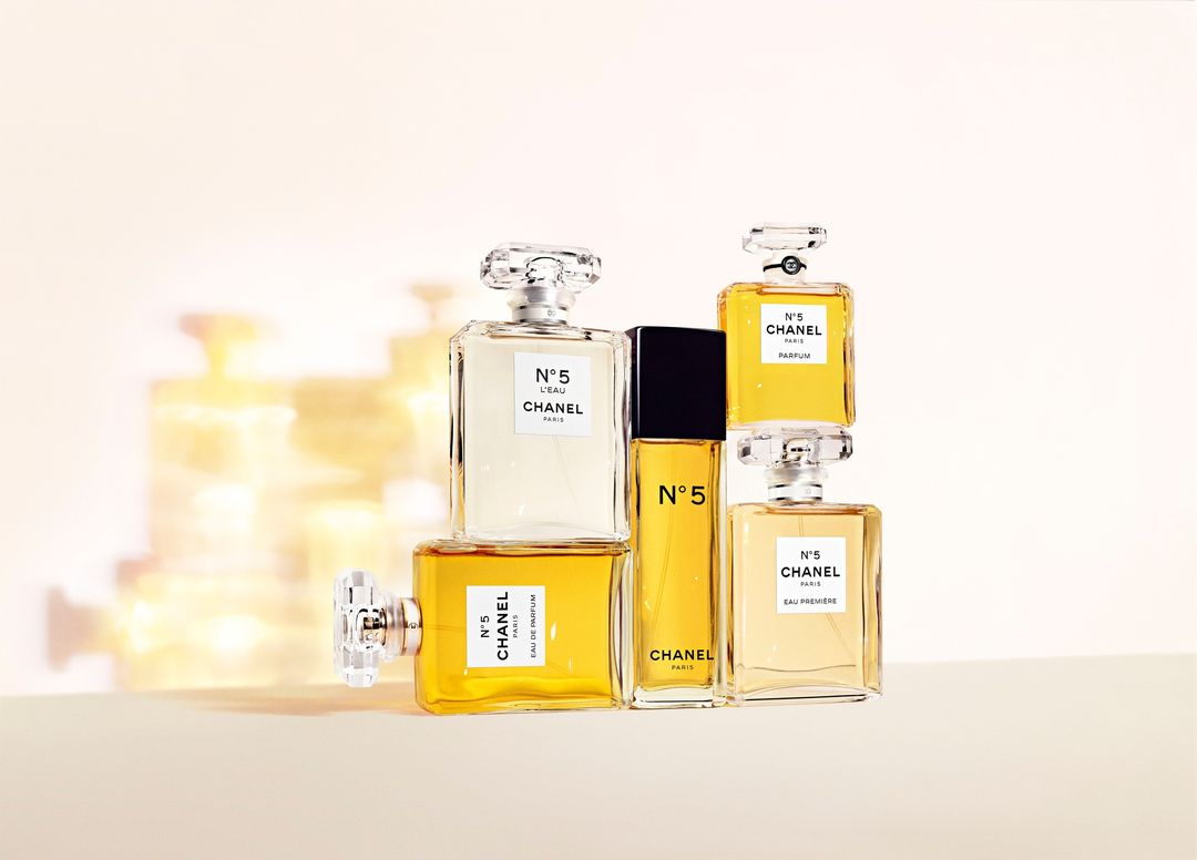 Several square Chanel No. 5 perfume bottles in clear and gold liquids are arranged artfully against a soft, light-toned backdrop.