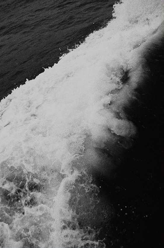 Black and white seascape features a powerful wave cresting and churning into foam. Ocean water is dark and textured.
