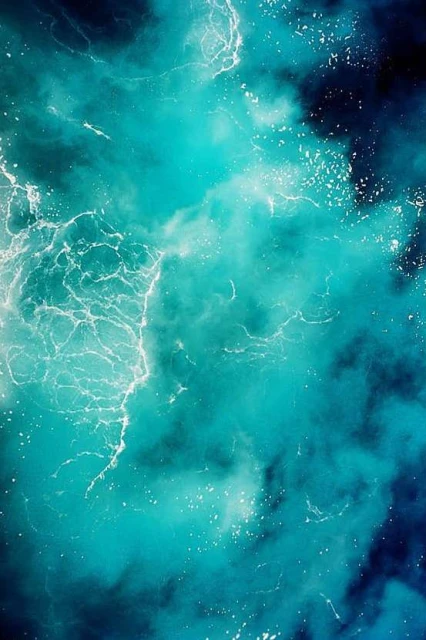 Aerial view of a turquoise ocean with white sea foam creating abstract patterns. The water transitions into dark blue.