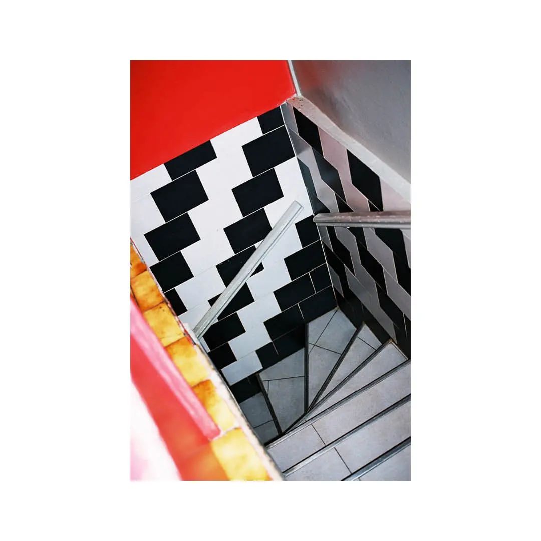 Angled shot of a stairway with gray stairs, silver handrail, and black and white geometric tile in an indoor location with red and gray walls.