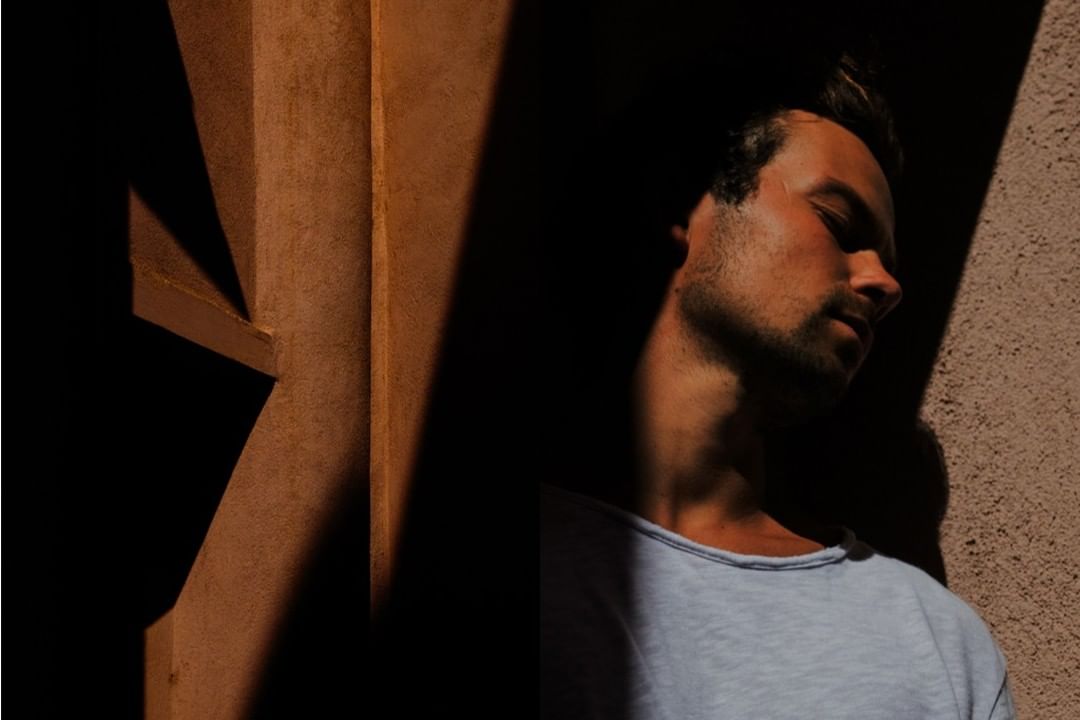 Portrait of a man with closed eyes, partially lit by sunlight, leaning against a brown textured wall with dark shadows.