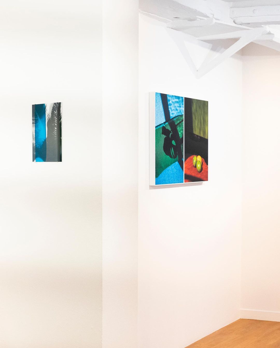Interior of a gallery featuring modern art pieces hanging on white walls, illuminated by bright light. Wooden floor visible.