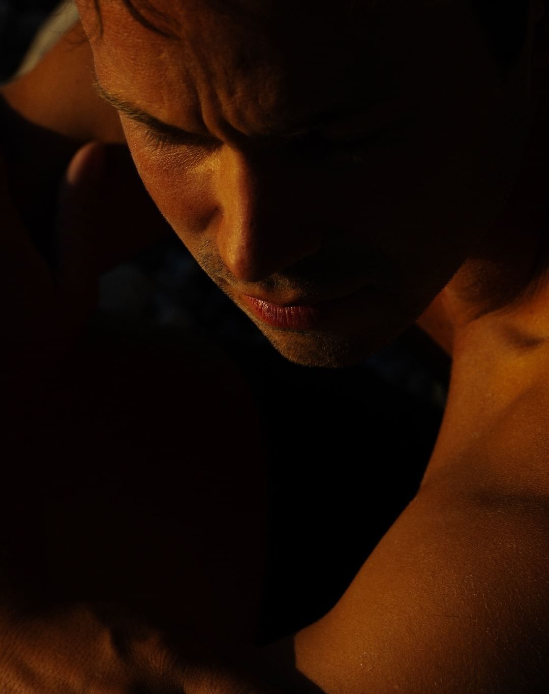 Close-up portrait of a shirtless adult male looking down, with strong shadows and warm lighting accentuating his face and upper torso.