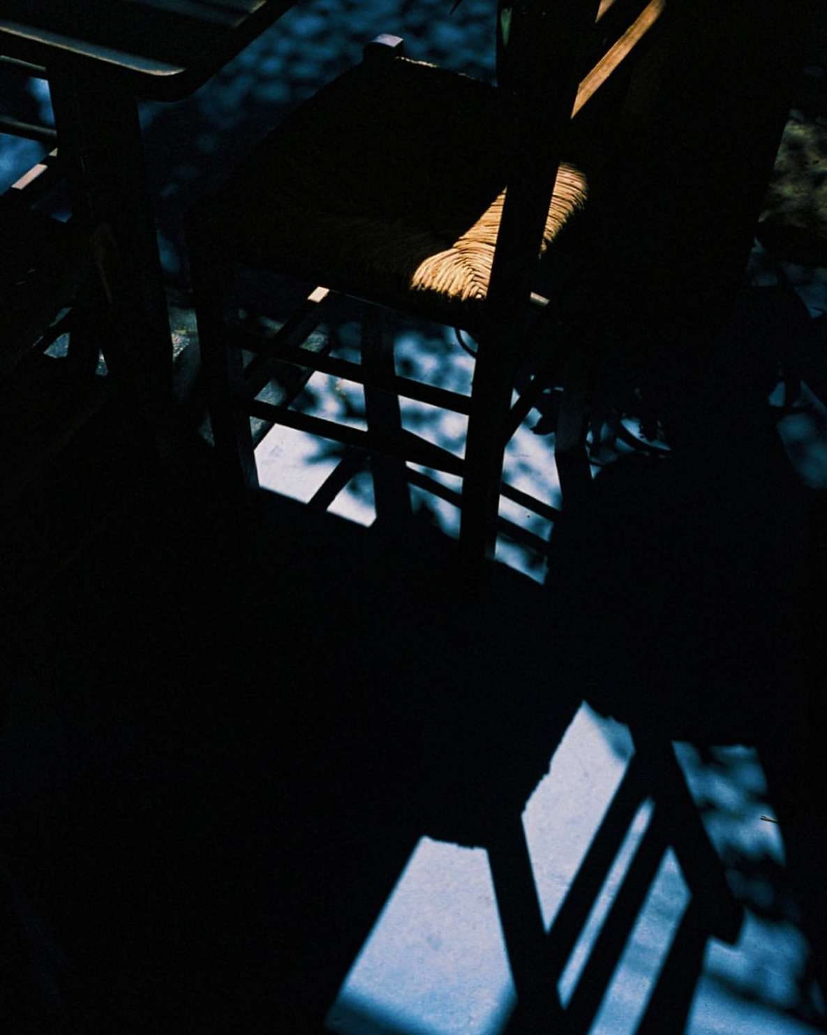 Abstract composition featuring a dark wooden chair with a woven seat, partially illuminated by a beam of sunlight creating long shadows.