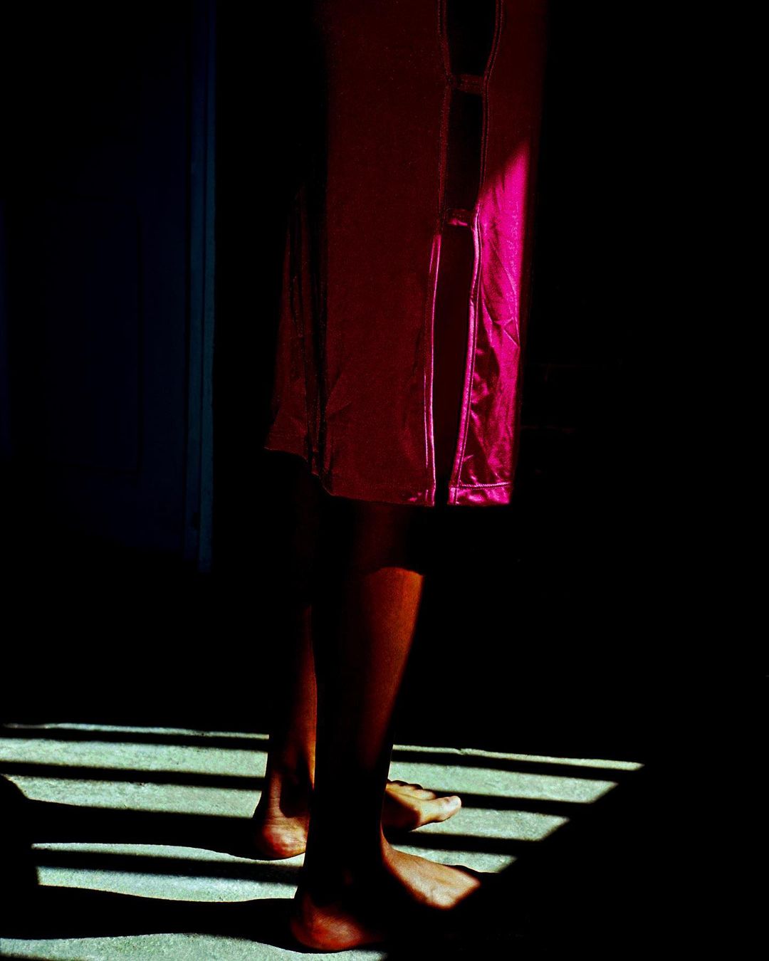 Lower portion of person in magenta gown standing in dark room with light stripes, shadows, and a blue door.