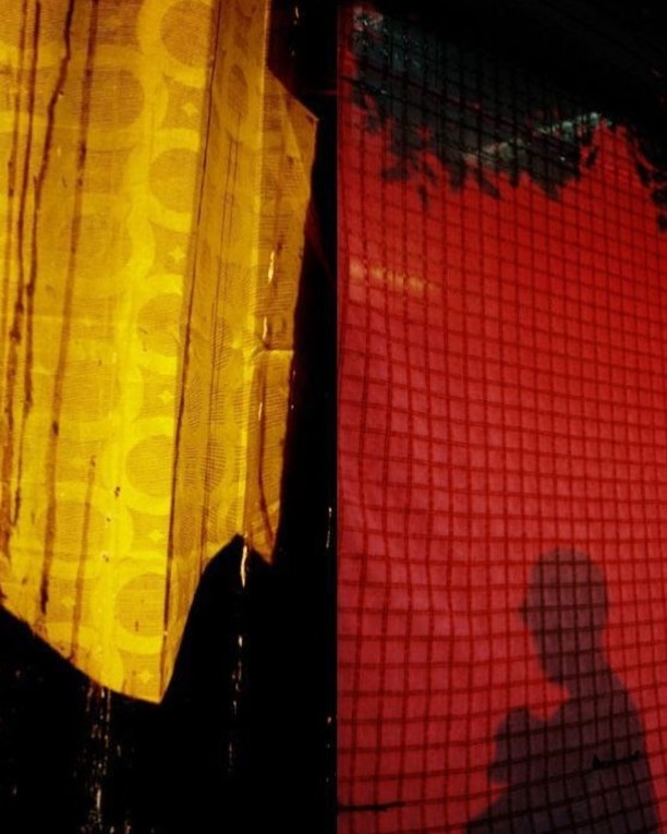 Abstract image of yellow and red window curtains with water droplets and a shadowy figure behind a red screen.