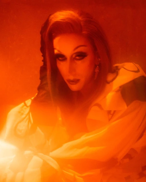 Dramatic portrait of a person with makeup and a red wig posing in an orange-lit photo studio, wearing large earrings, and a gown.