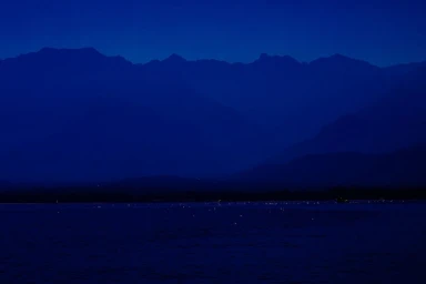 Deep blue nighttime landscape showing mountain silhouettes across a dark body of water with distant lights.