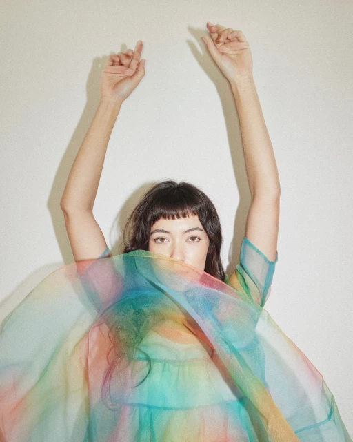 A young woman with dark hair and bangs raises her arms above her head, partially obscuring her face with a colorful, translucent fabric.