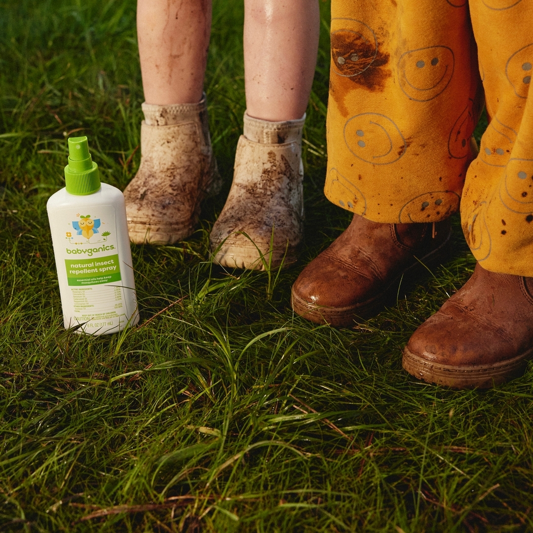 Close-up of Babyganics insect repellent next to children's dirty boots and pants with smiley-face prints on green grass.