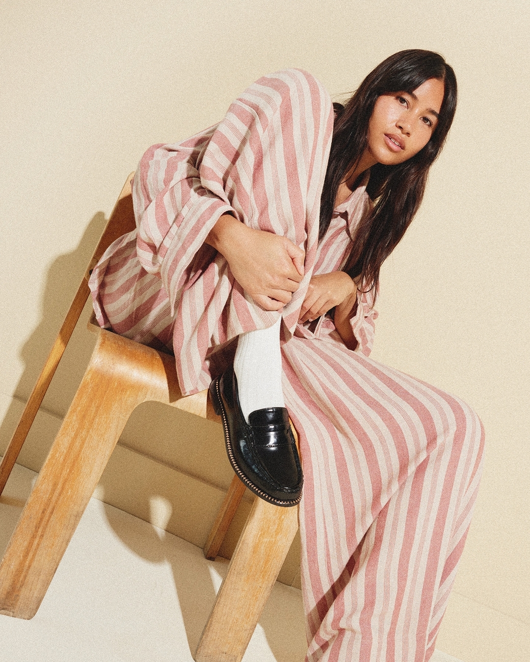 A young adult female poses sitting on a wooden chair wearing a striped pajama set with glossy black loafers against a cream backdrop.