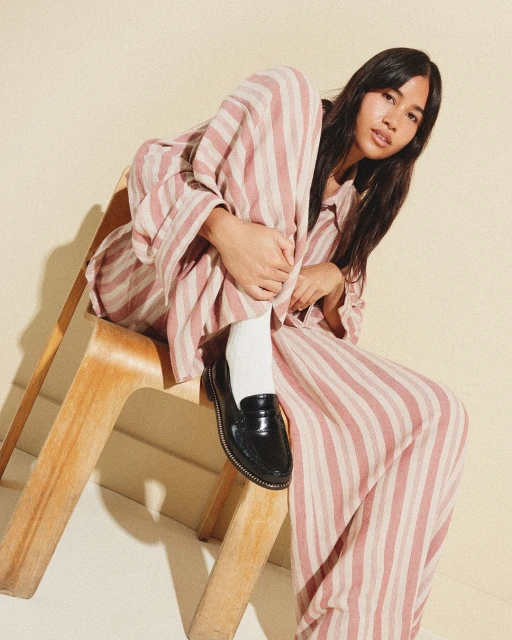 A young adult female poses sitting on a wooden chair wearing a striped pajama set with glossy black loafers against a cream backdrop.