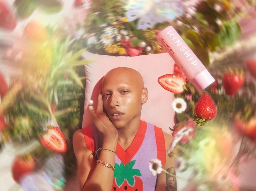 A person with a shaved head poses with a Kinship product, strawberries, and daisies against a blurred floral backdrop and pink pillow.