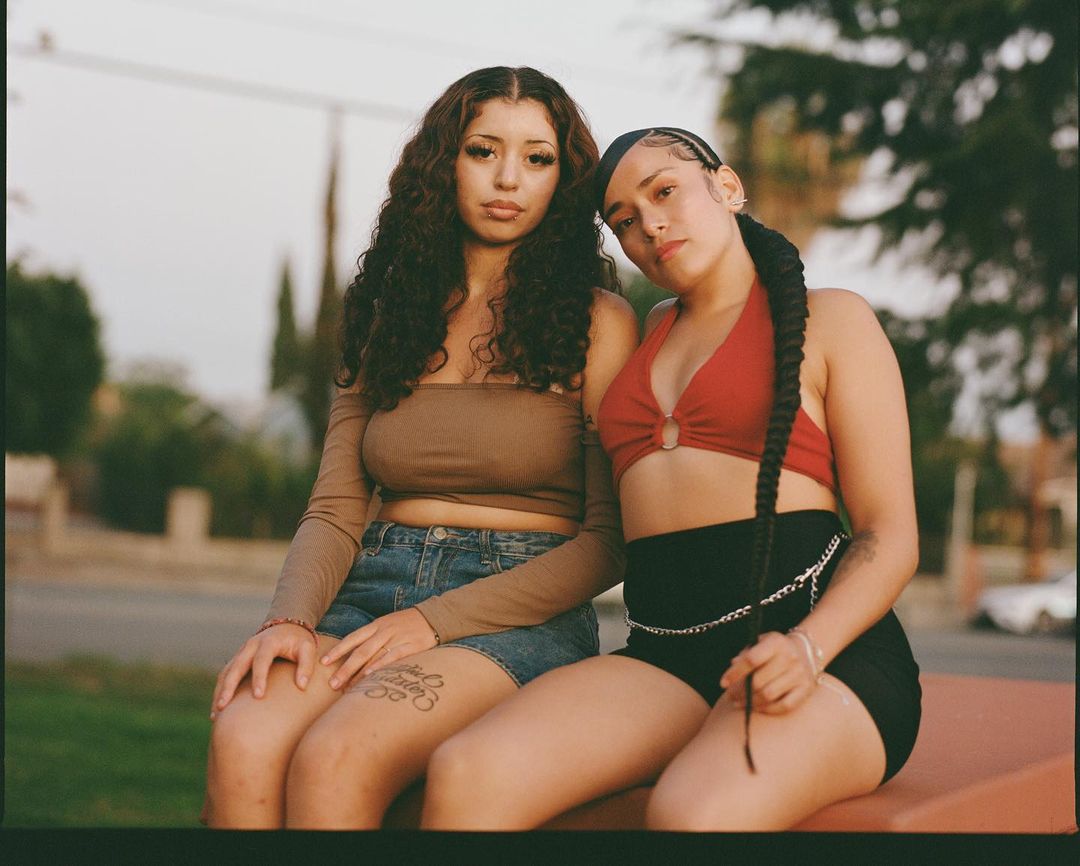 Two young women pose outdoors in warm sunlight. The woman on the right has a long braid and a red halter top. The other has long, curly hair.