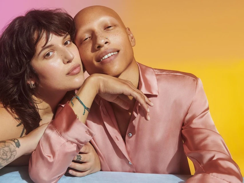 A close-up studio shot of two people, one bald and one with dark hair, in pink button-down shirts against a vibrant yellow to pink gradient background.