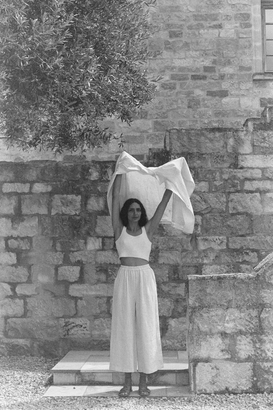 Woman in white separates holds a sheet in front of a stone wall with tree branches in the upper-left corner on a sunny day.
