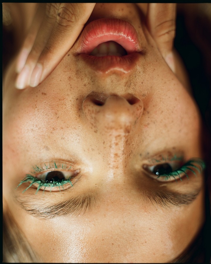 Close-up portrait of a fair-skinned woman with freckles, green mascara, and pink lips, angled with hand near her face.