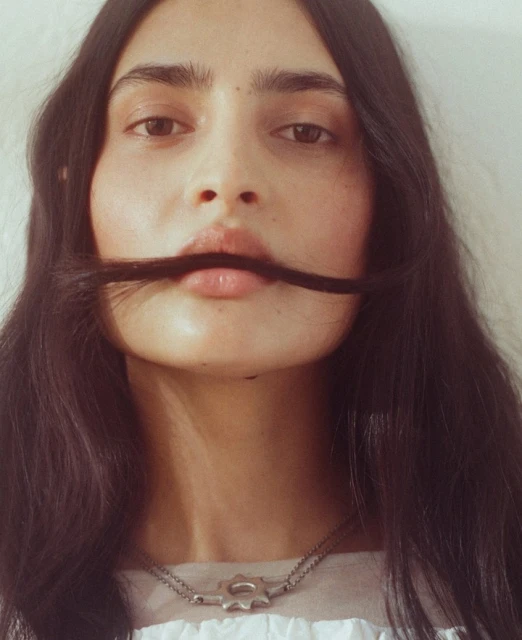 Portrait of a long-haired woman holding her hair over her upper lip, creating a mustache effect, wearing a pendant necklace, indoors.
