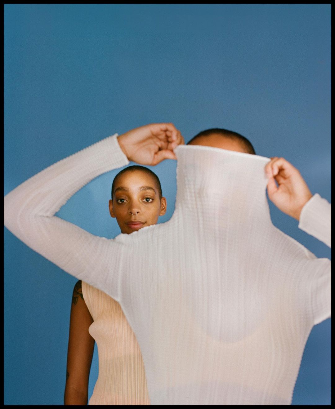 Two women pose in a photo studio against a blue backdrop. One woman covers her face with a sheer white turtleneck.