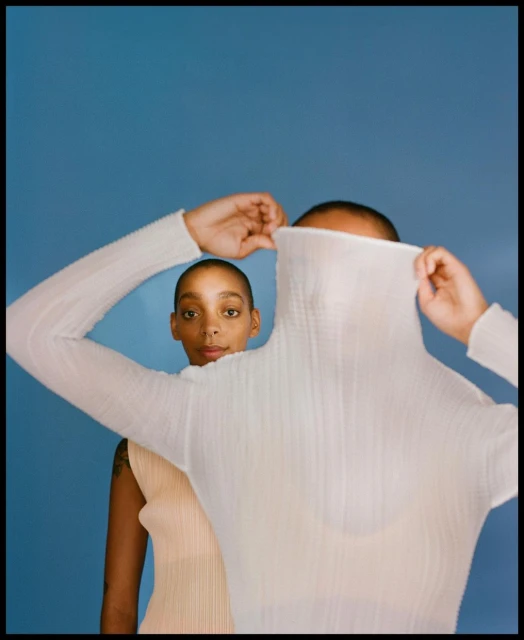 Two women pose in a photo studio against a blue backdrop. One woman covers her face with a sheer white turtleneck.