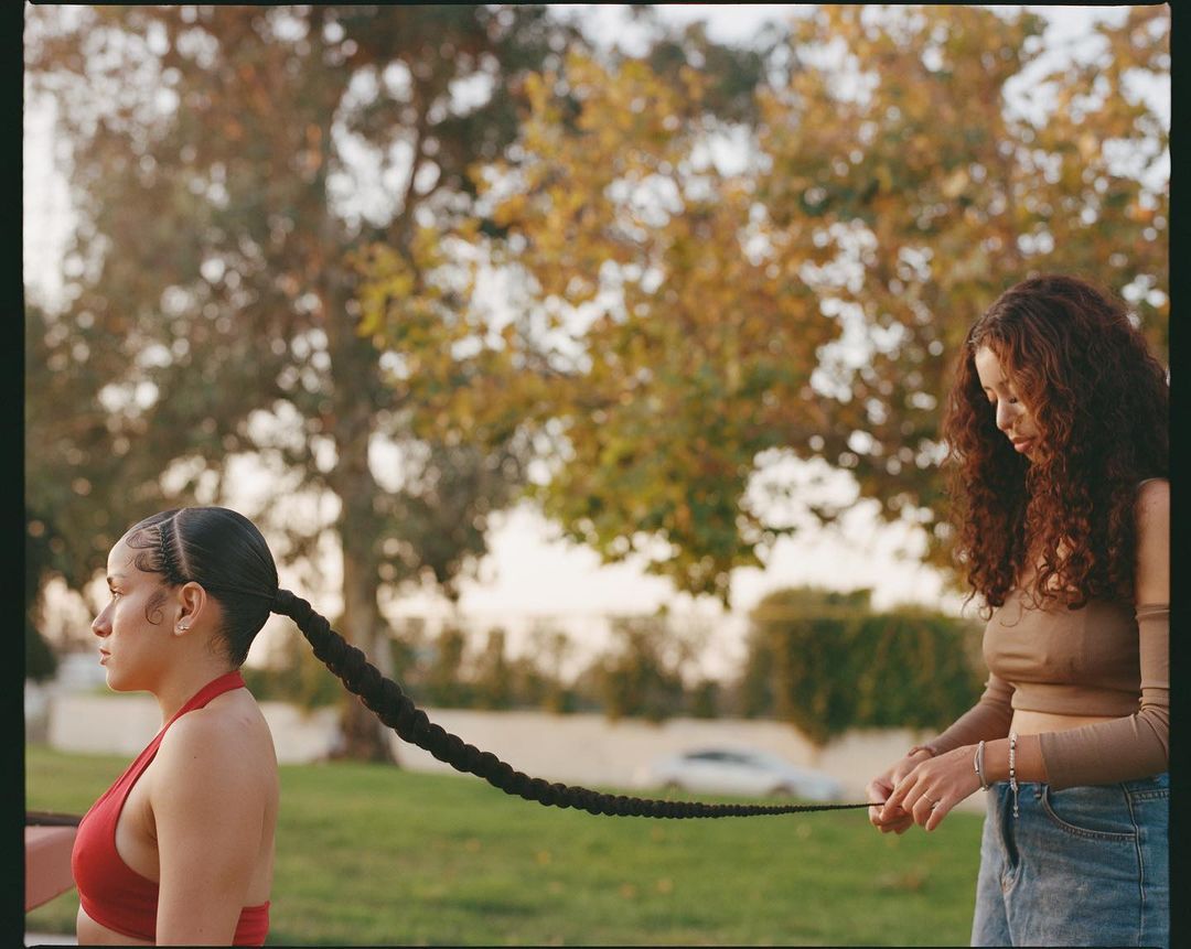 Two women outdoors, one braiding the other's long, dark hair in a park setting with trees and greenery; warm, natural lighting.