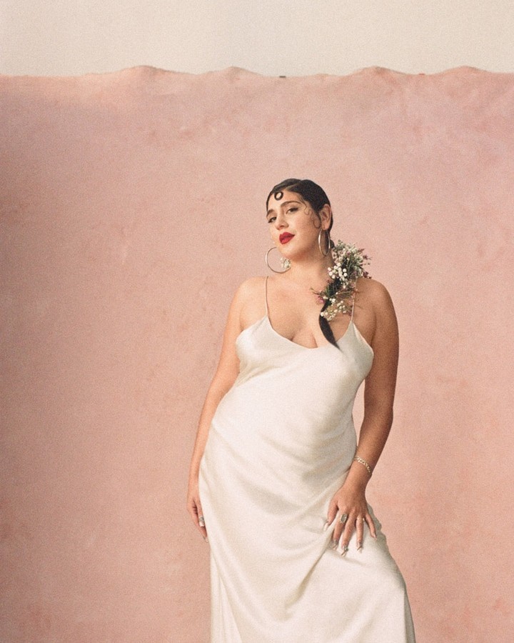 Elegant woman in white dress poses in front of a soft pink wall, adorned with delicate white flowers in her hair, creating a serene scene.