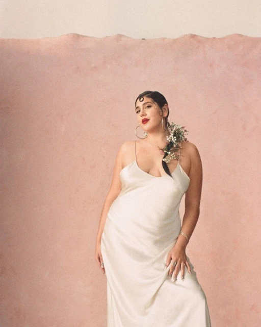Elegant woman in white dress poses in front of a soft pink wall, adorned with delicate white flowers in her hair, creating a serene scene.