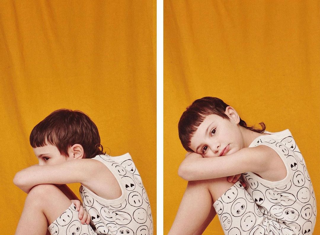 Child in patterned outfit sits in front of a yellow background, captured in a studio shot with medium framing and portrait orientation.