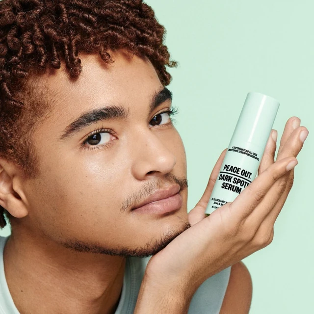 Close-up studio shot of an adult man holding a skincare serum bottle against a light green backdrop, showcasing the product.