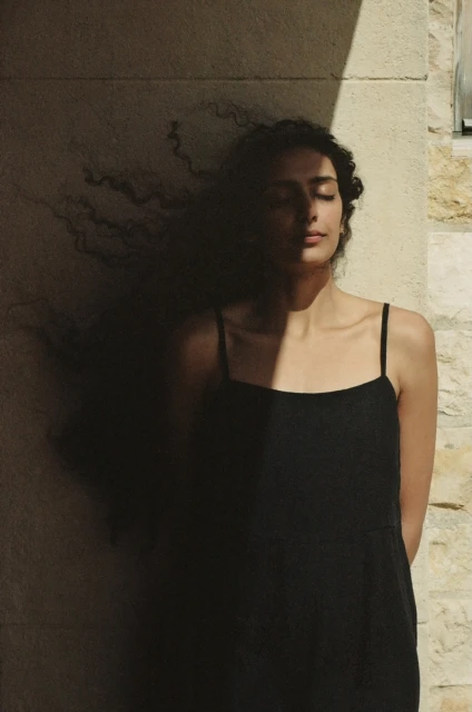 Woman with curly hair in black dress standing against stone wall with light and shadow in outdoor setting with closed eyes.